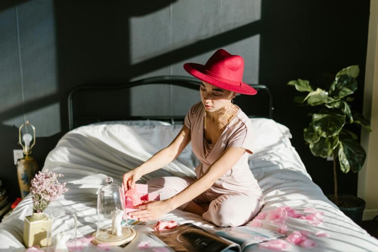 A woman wearing a red hat sits on a bed, engaged in crafting with pink flowers.